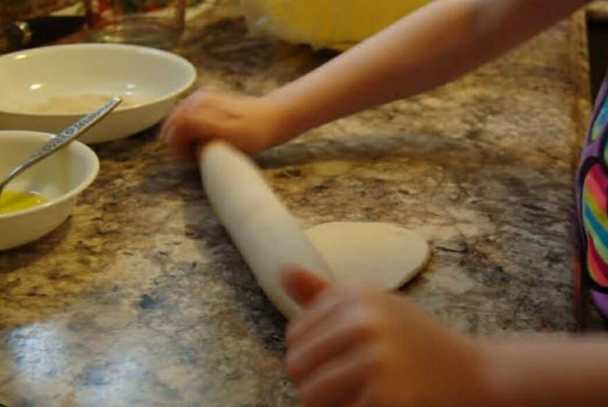 Child's hands using a rolling pin to roll out roti on a stone benchtop.