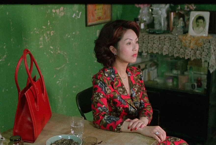 Scene from the 2004 Hong Kong horror film Dumplings, of a woman with short brown hair sitting at a wooden table indoors, with a large red tote bag visible to her left.