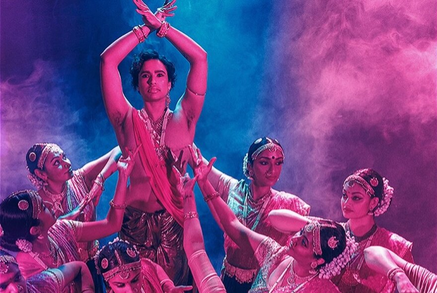 Indian dancers in traditional attire dancing on a pink and blue-lit stage.