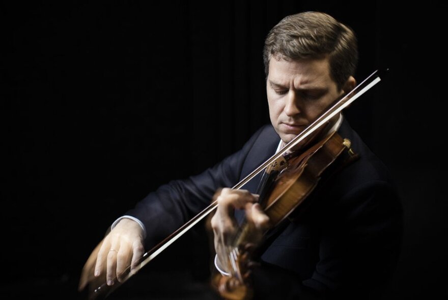 Classical violinist wearing a black suit, looking down at his instrument as his hand holds the bow.