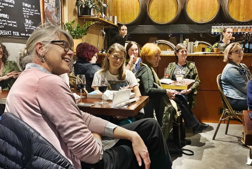 People seated at tables in a wine bar with barrels, listening and smiling.