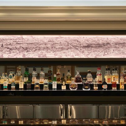 An illuminated back bar featuring multiple tiers of liquor and spirit bottles, topped by a long, backlit panoramic sketch of a historic cityscape.