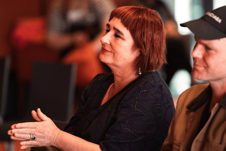 A woman in a black shirt with dark red hair clapping her hands, seated next to a man in a black baseball cap and brown jacket.