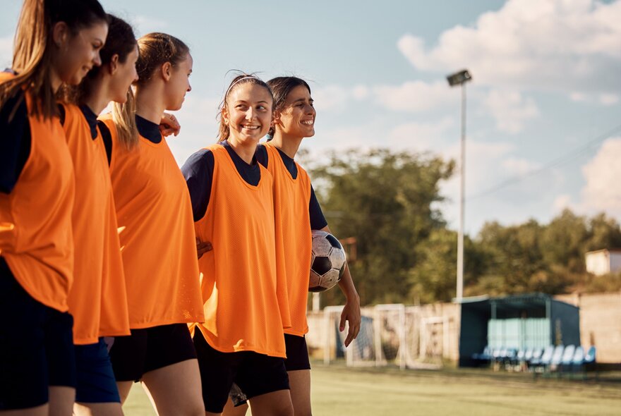 A smiling group of 5 young women in orange bibs on a sporting field. 