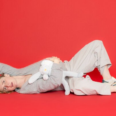 A performer lying on the ground, clutching her belly and holding a soft toy, against a red background.