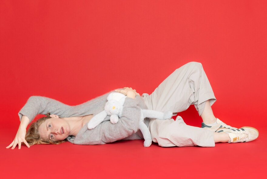 A performer lying on the ground, clutching her belly and holding a soft toy, against a red background.