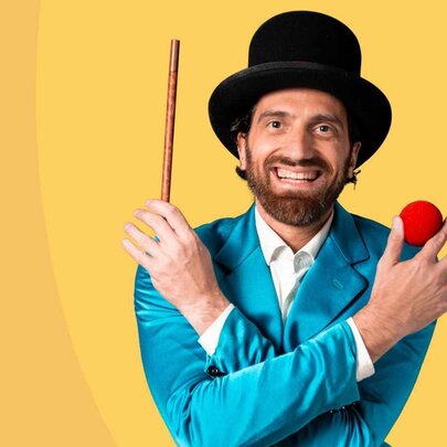 The Great Armando, a children's magician wearing a black top hat, a turquoise suit, holding a short wand in one hand and four red balls between his fingers in the other hand.