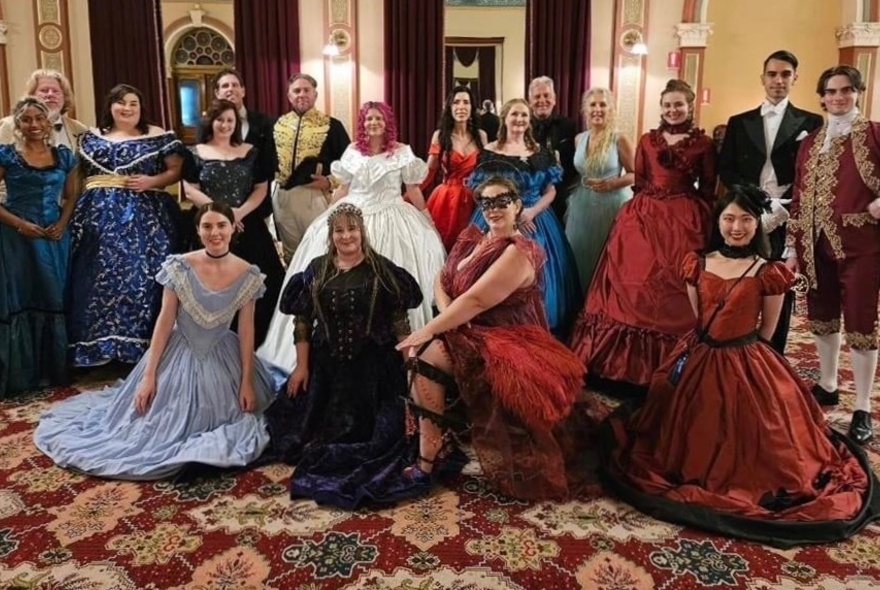 A group of people wearing period costumes seated and standing in an ornate room.