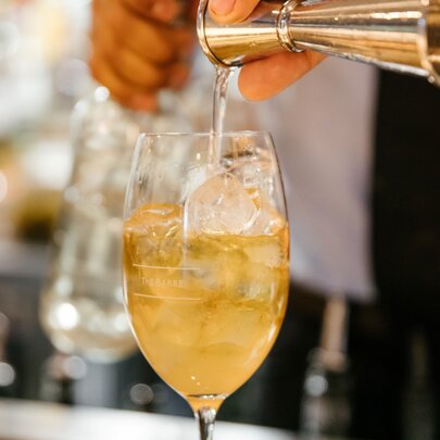 A shot of clear spirit being poured into a yellow-toned cocktail in a wine glass.