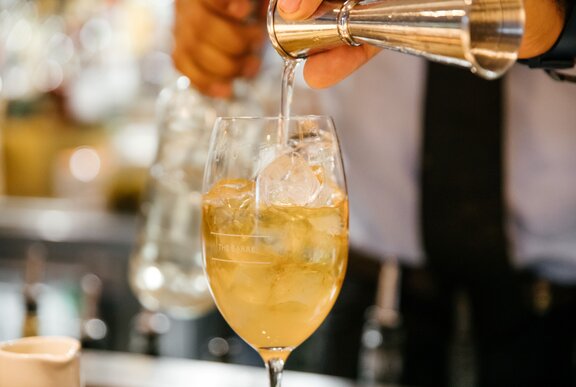 A shot of clear spirit being poured into a yellow-toned cocktail in a wine glass.
