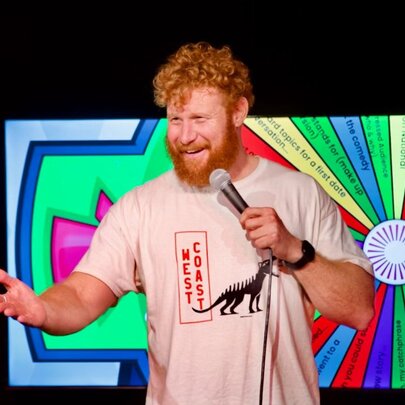 Comedian holding a microphone with his hand outstretched, in front of a screen of a spinning wheel.