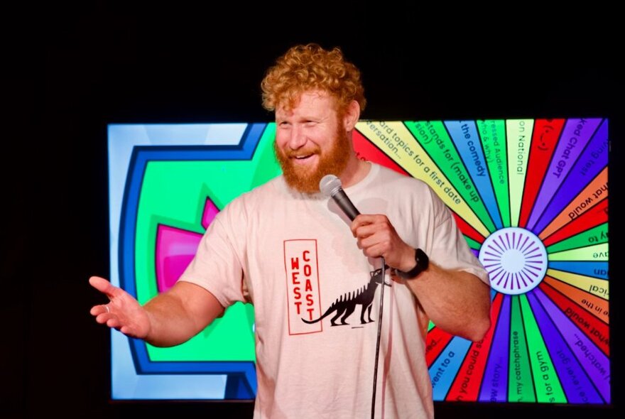 Comedian holding a microphone with his hand outstretched, in front of a screen of a spinning wheel.