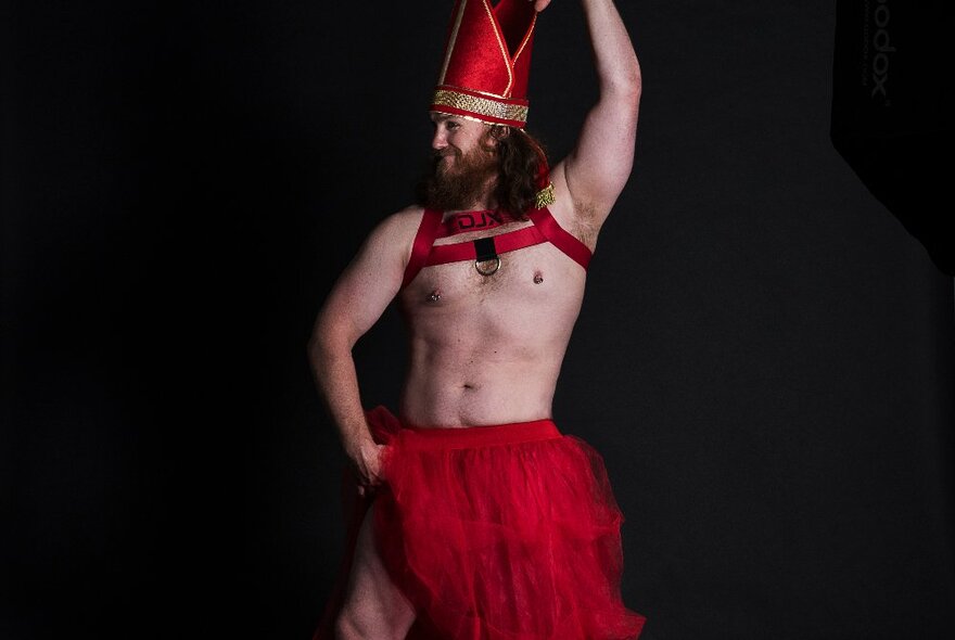 A bearded performer wearing a red tulle skirt, red chest straps on his naked torso, and a red bishop's hat, standing in profile against a black background.