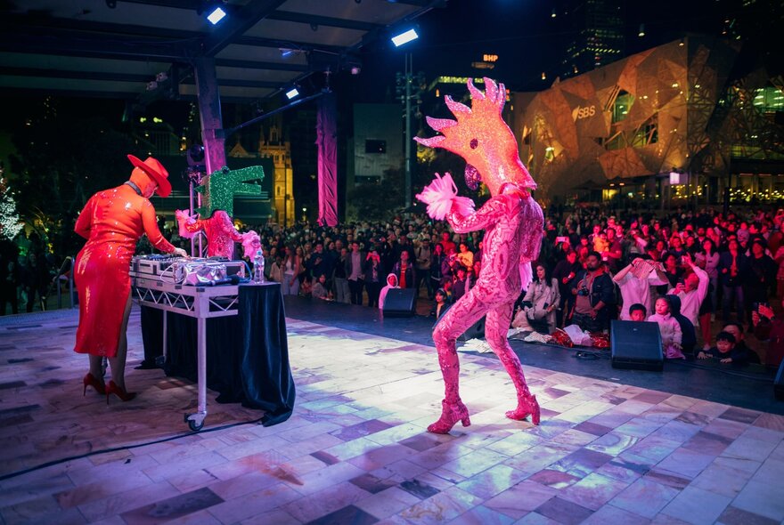 The main stage at Fed Square at night, with a DJ at a mixing booth and a person in a pink body stocking and elaborate headdress on stage, performing to a large crowd in the plaza.