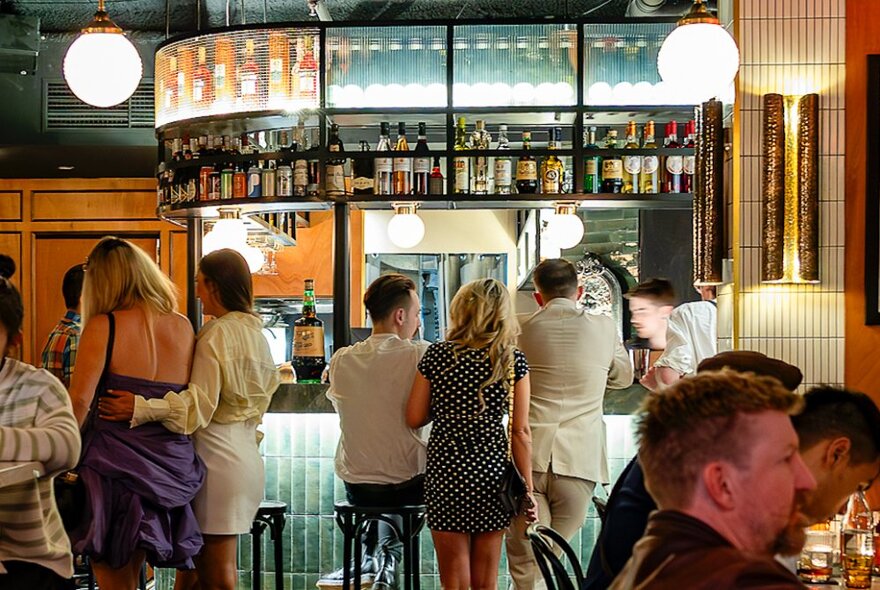 People socialising and standing at a bar, some seated on high bar stools.