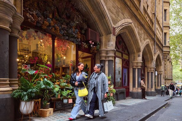 Two women walking past a florist with shopping bags and a bunch of flowers in their arms.