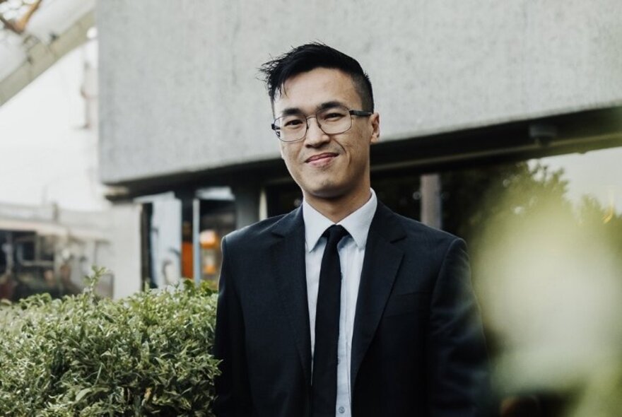 Author Siang Lu, wearing a white shirt and tie, a dark suit jacket and posed outside among shrubs with a building behind him.