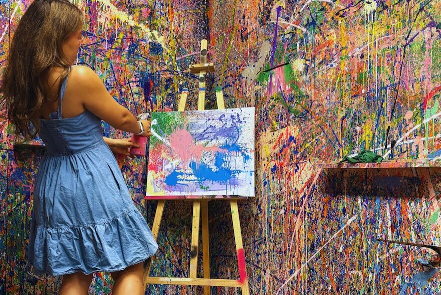 A child holding a paintgun, squirting paint on a canvas in a room with walls covered in paint splashes.