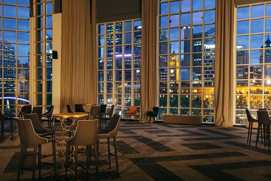 An empty room with tall windows, patterned carpet and widely-spaced tables and chairs; Melbourne's nighttime skyline seen through the windows.
