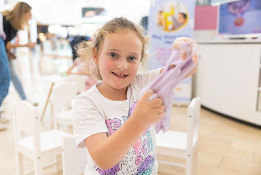 A smiling young girl stretching slime in a room with small tables and chairs set up for an activity.