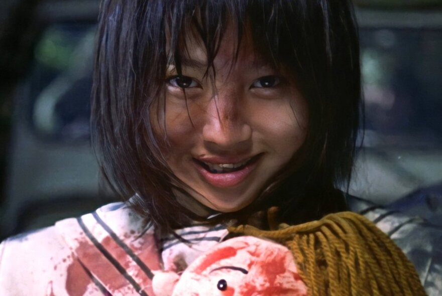 Still from a Japanese horror film of a face of a young girl smiling in a creepy way, her top is blood splattered.