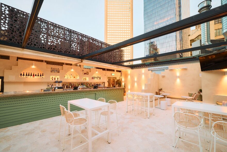 Rooftop bar with white paving, white seating and tables, green-tiled bar with city buildings in the background.