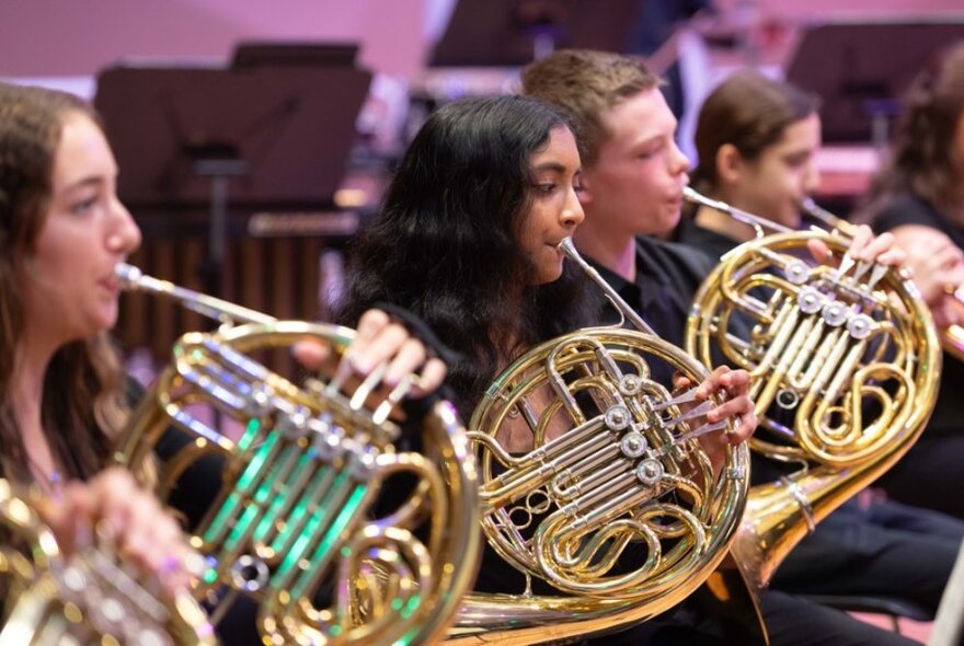 Three young French horn musicians dressed in black, seated on stage.