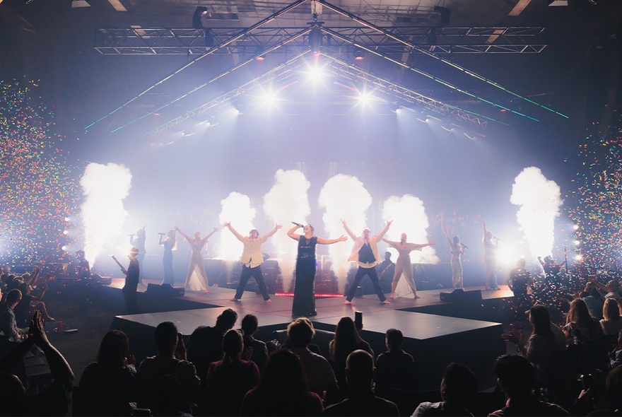 A singer in a long dark dress, with dancers behind her, performing a show on the stage at Festival Hall to a large audience.