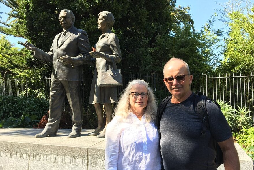 Two people on a walking tour standing in front of a bronze state in a public park in East Melbourne.