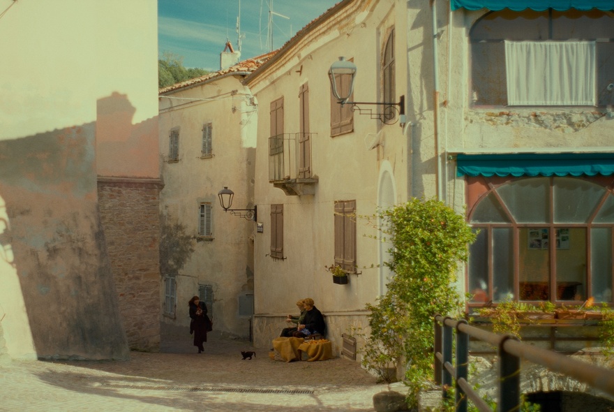 A still image from a film showing a small Italian streetscape with people sitting on boxes.