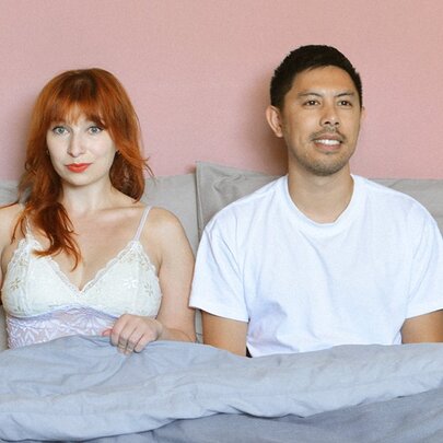 A man and a woman sitting up in bed, he is smiling and she's looking slightly bewildered. 