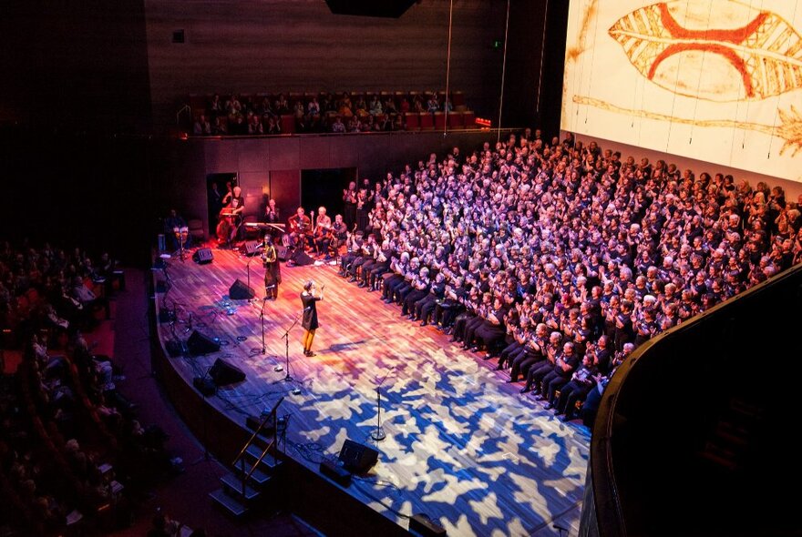 Conductor leading massed rows of choir singers on stage.