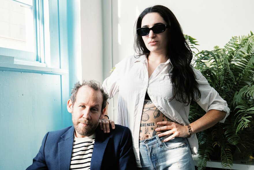 Singers Ben Lee (seated) and Georgia Maq standing beside him with her hand on her hip and sunglasses on.