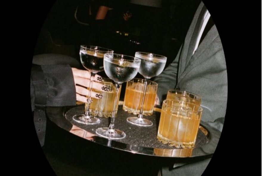 A person in a grey suit carrying a silver tray of cocktails.