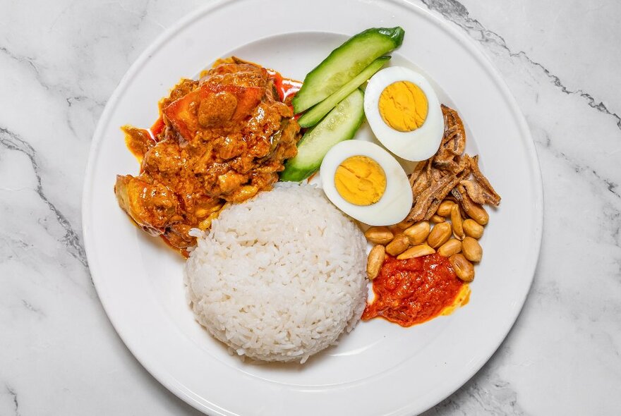 Looking down at a plate of nasi lemak  - rice, boiled eggs, cucumber, nuts and shrimp.