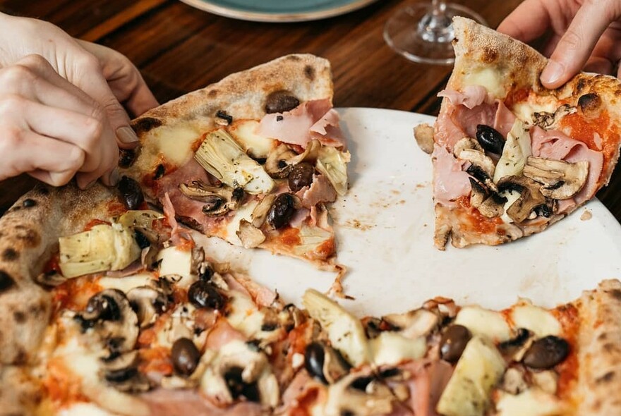 Hands taking slices of artichoke pizza from a plate.