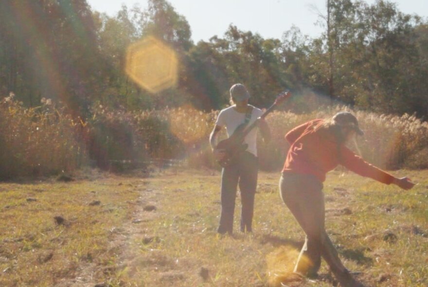 Two people in a field outdoors, one of them playing electric guitar and the other person moving as if dancing.