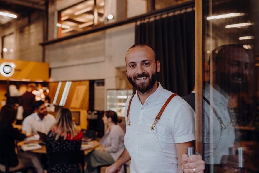 A man wearing a white apron, holding open the glass door to a coffee shop, with a welcoming smile.