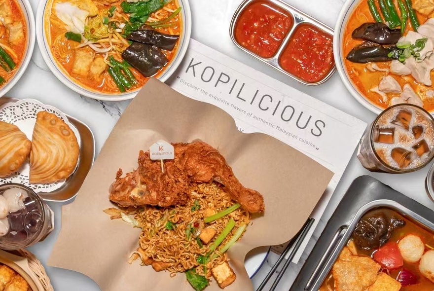 A variety of delicious Malaysian hawker-style streetfood in bowls surrounding fried chicken and noodles resting on a piece of brown paper.