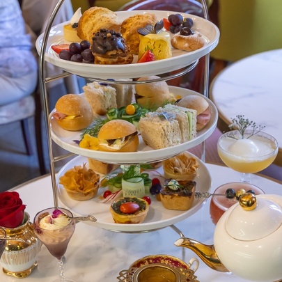 A three-tiered high-tea offering with cakes and small savoury bites, someone pouring tea from a pot. 