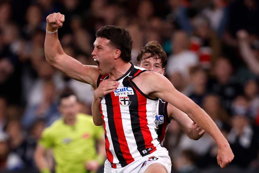 Two St Kilda AFL players running and cheering, with a blurred stadium crowd in the background.