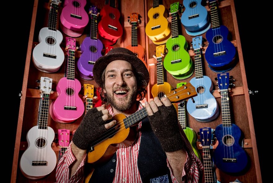 A smiling man with a beard wearing a hat, playing a ukulele, in front of a wall of brightly coloured ukuleles.