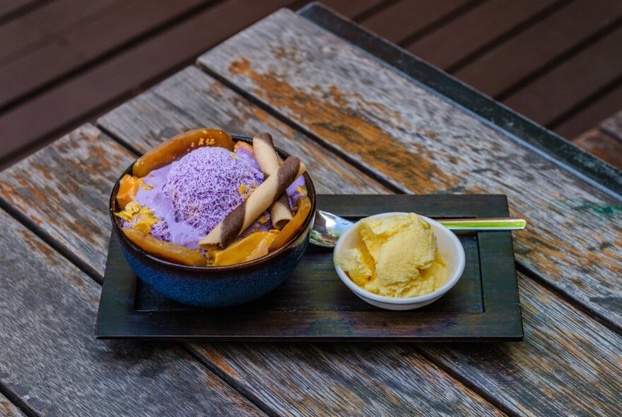 purple shaved ice in a bowl topped with other ingredients alongside a scoop of ice cream on a wooden table.