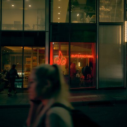 Exterior of Supernormal restaurant on a winter's night, with pedestrians walking past, the view though the window is of a warm inviting space. 