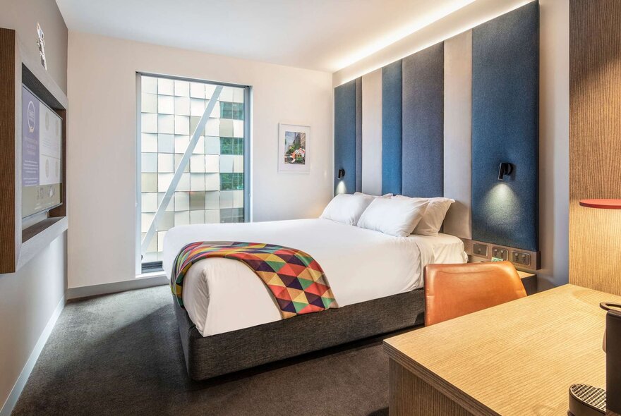 A contemporary hotel room at the ibis Styles Melbourne Southbank featuring a queen-sized bed with a white duvet and a colourful, patterned throw blanket.