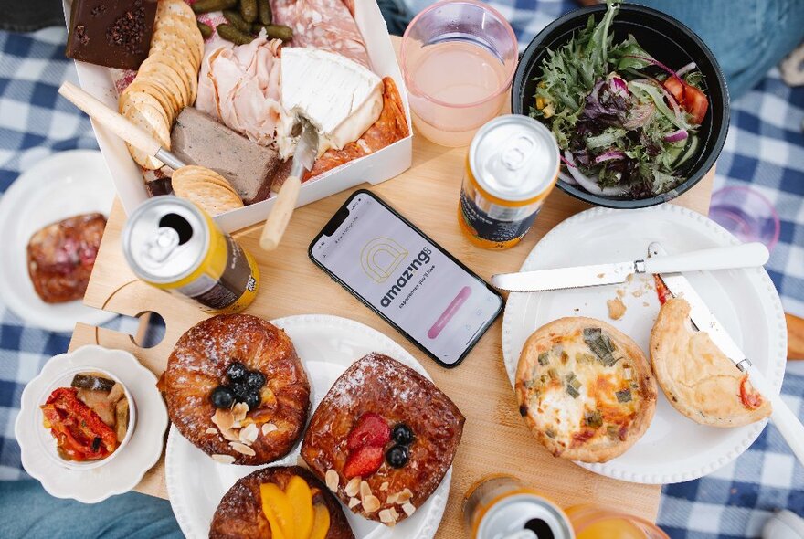 Picnic spread of dishes, boxed food and drinks on a blue gingham tablecloth.