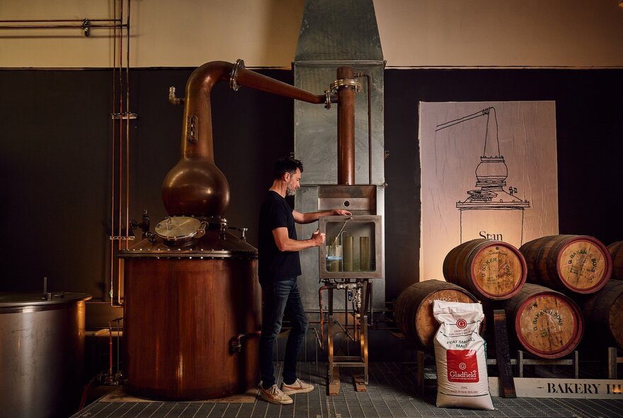 A man stands next to a large copper still and wooden barrels in a distillery.