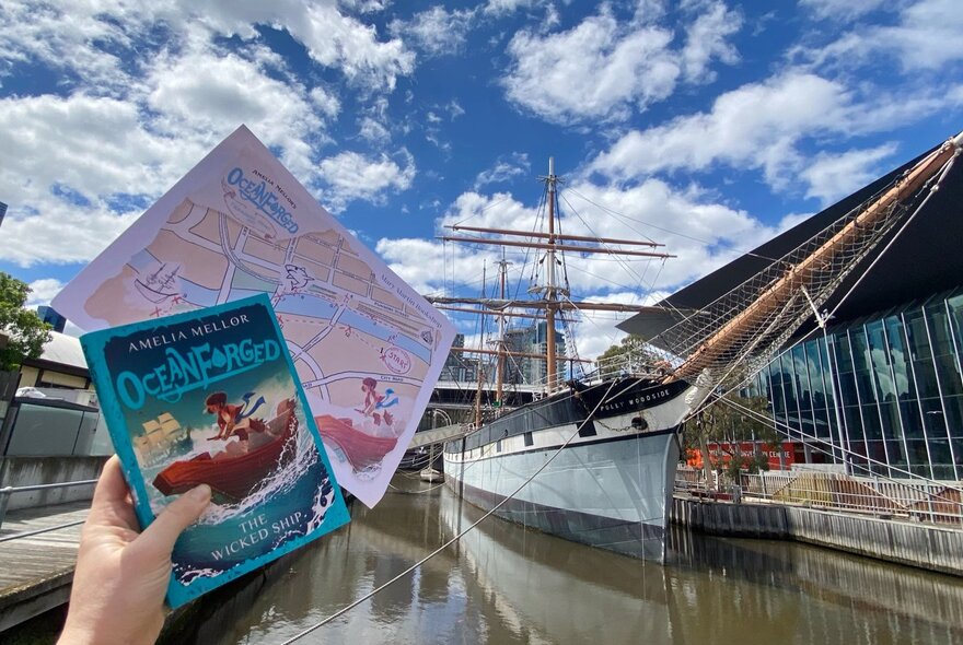 A book and treasure map held up by a person's hand in front of the Polly Woodside tallship in Melbourne.