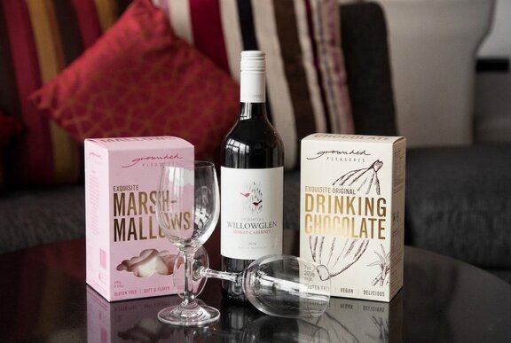 One pink and one white box of premium drinking chocolate either side of a small wine bottle with two empty wine glasses in front of them, all displayed on a black surface.