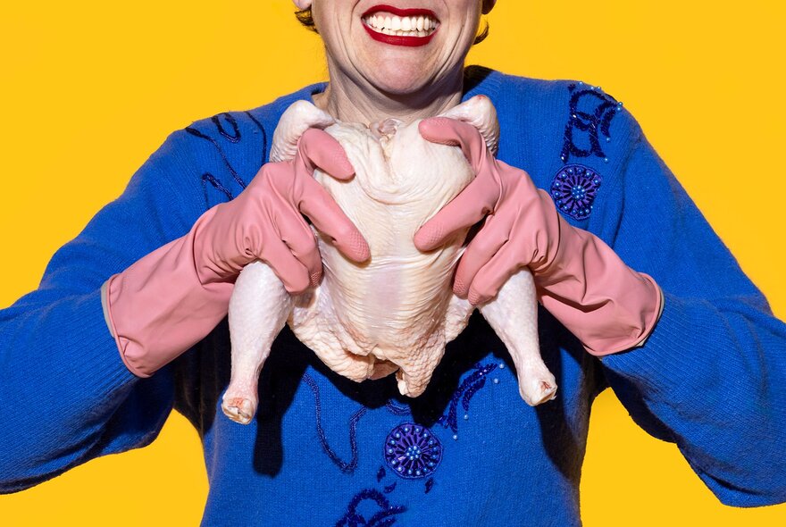A woman wearing a blue embellished cardigan and pink rubber gloves, holding a raw chicken carcass as though she's going to pull it apart; yellow background.